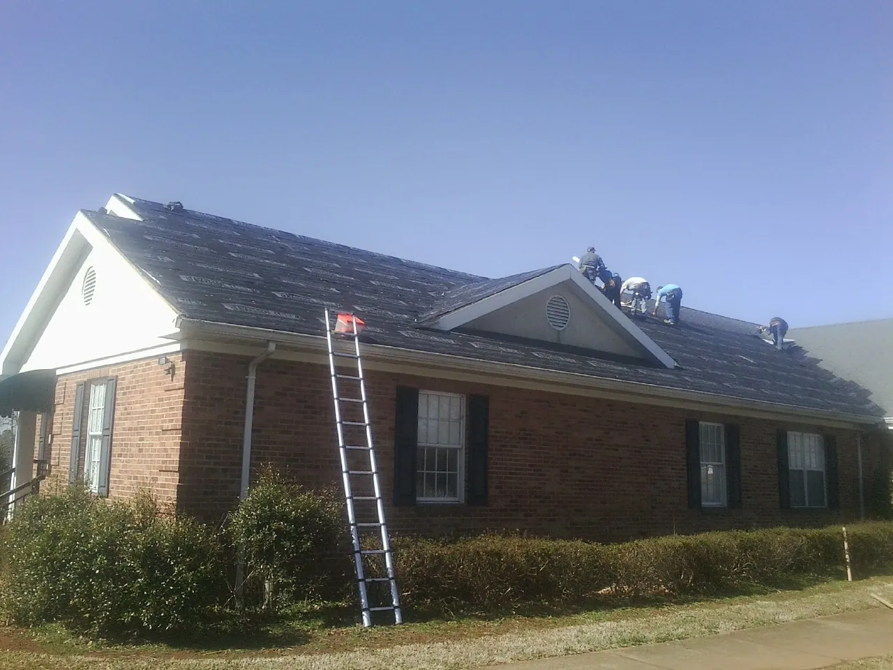 Crew tearing shingles off a brick ranch roof with a ladder set — Emergency Roof Repair work by our crew in Kerkhoven