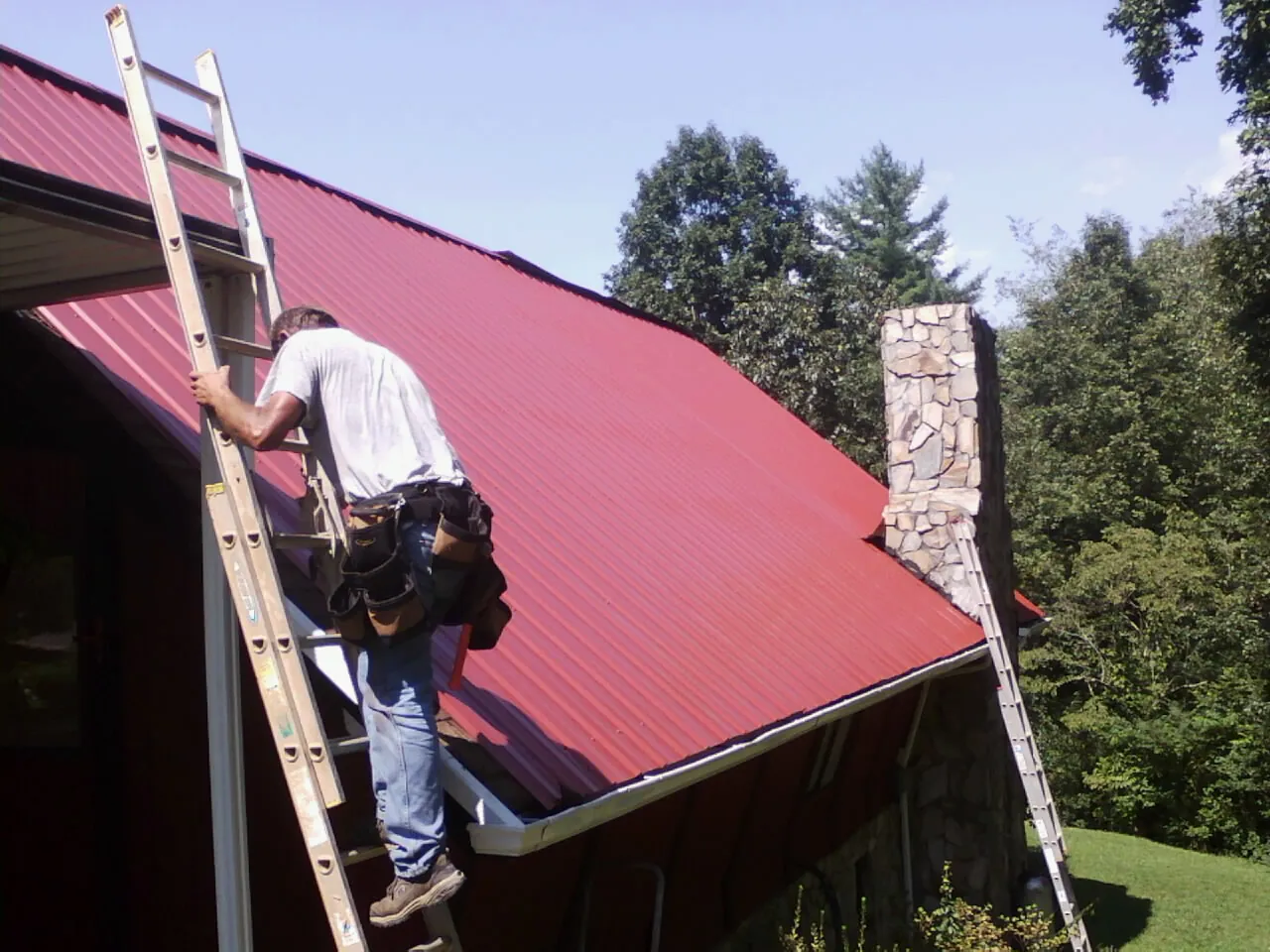 Roofer on a ladder beside a red metal roof and stone chimney — Soffit & Fascia Repair by Roofing Brothers in Kerkhoven